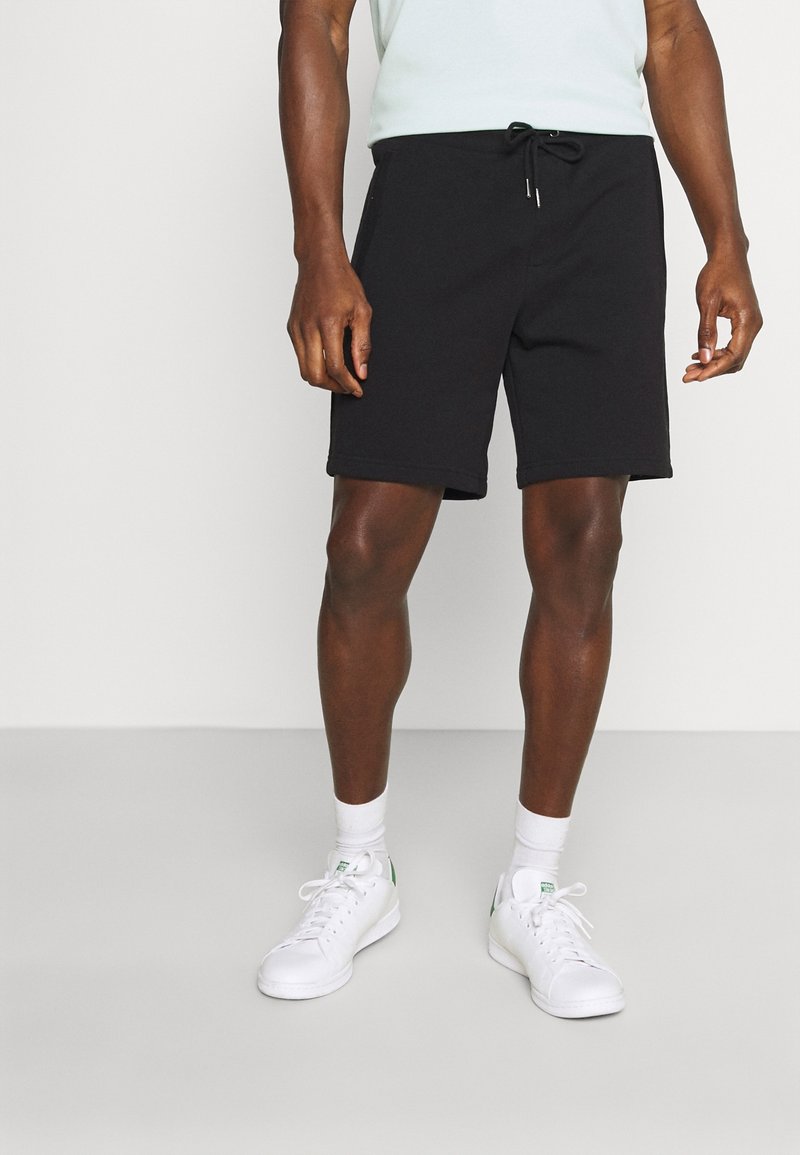 Shorts de algodón negros con cintura ajustable, corte a la rodilla y bolsillos laterales, combinados con zapatillas blancas y calcetines hasta el tobillo.