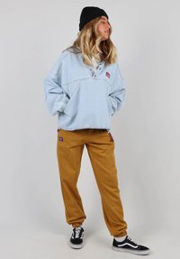 Pull bleu clair en denim avec un col et des boutons sur le devant, associé à un jogging moutarde avec des poches. Des baskets noires complètent la tenue.