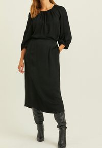 Schwarzes Midikleid mit rundem Ausschnitt, puffartigen Ärmeln und lockerer Passform. Trägt kniehohe schwarze Stiefel, die Textur und geschichtete Interessen hinzufügen.
