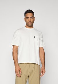 T-shirt bianco in cotone con scollo rotondo, maniche corte e taschino sul lato sinistro del petto. Presenta un piccolo dettaglio del logo nero. Indossato con pantaloni corti beige.