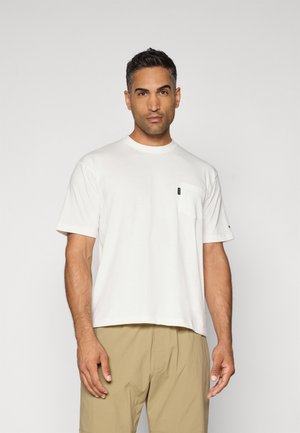 T-shirt en coton blanc avec un col rond, des manches courtes et une poche sur la poitrine gauche. Présente un petit logo noir. Porté avec un short beige.