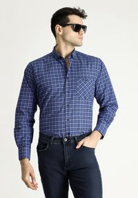 Camicia a quadretti blu con maniche lunghe e taschino sul petto, abbinata a jeans scuri. Occhiali da sole indossati, capelli acconciati.