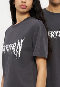 Δύο άτομα φορούν σκουρόγριες μπλούζες με λευκό γραφικό κείμενο, μια γυναίκα μπροστά με μακριά κατσαρά μαλλιά και ασημένια σκουλαρίκια κρίκους.