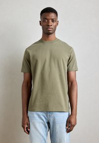 Olive green cotton t-shirt with short sleeves, round neck, and a relaxed fit, paired with light blue jeans. Minimalist design, no visible patterns.