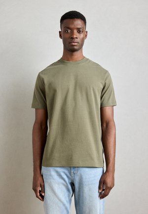 Olive green cotton t-shirt with short sleeves, round neck, and a relaxed fit, paired with light blue jeans. Minimalist design, no visible patterns.