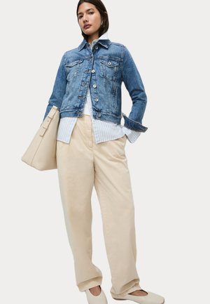 Chaqueta de mezclilla con cierre de botones, color azul claro, cuatro bolsillos. Combinada con una camisa a rayas y pantalones anchos beiges. Bolso beige.