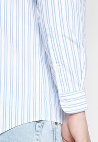 Camicia a maniche lunghe a righe verticali blu chiaro e bianco, con il polsino abbottonato sul polso di una persona, sopra jeans in denim blu chiaro.