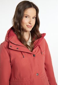 Giacca isolante rossa con cappuccio, dotata di chiusura con zip e bottoni, coulisse e texture liscia, esposta su un modello contro uno sfondo bianco.