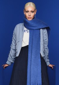 Hellblau Bomberjacke mit Raffung, cremiges Hemd, dunkler plissierter Rock und ein langer blauer Schal, vor einem blauen Hintergrund.
