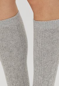 Detailný záber nôh v svetlošedých pletených kolenných ponožkách s textúrovaným vzorom a rebrovanými manžetami.