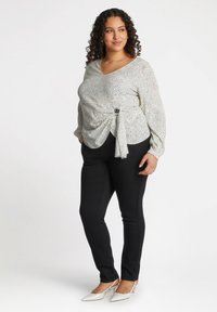 Weiße Bluse mit schwarzen Punkten, V-Ausschnitt, langen Ärmeln, gerafftem Vorderteil mit Knotendetail; kombiniert mit enganliegenden schwarzen Hosen und weißen Absätzen.