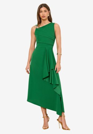 Frau in ärmellosem asymmetrischem grünem Kleid mit Rüschendetail, goldener Brosche auf der Schulter, goldenem Armband und goldenen Sandaletten mit Absatz.