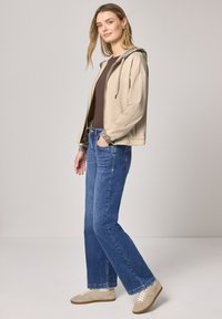 Kvinde stående iført beige hættetrøje med lynlås og leopardprintkant, brun skjorte, blå jeans og beige sneakers på lys grå baggrund.