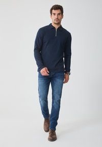 Polo de manga longa azul marinho com gola de fecho éclair, combinado com jeans azuis. Botas castanhas de atacador completam o visual; tecido liso com um acabamento texturizado.