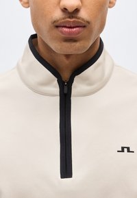 Κοντινό πλάνο ενός ατόμου που φοράει μπεζ πουλόβερ με μαύρο φερμουάρ και μικρό μαύρο γεωμετρικό λογότυπο στο στήθος.