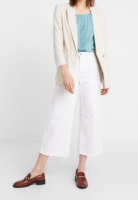 Blazer en lin beige, haut en dentelle bleu sarcelle, jean large blanc raccourci, mocassins marron avec accents métalliques. Lignes épurées et matériaux texturés.