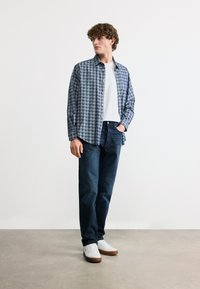 Νεαρός άνδρας στέκεται με το χέρι στην τσέπη φορώντας μπλε πουκάμισο με καρό, λευκή μπλούζα, σκούρο τζιν και λευκά αθλητικά παπούτσια σε απλό φόντο.