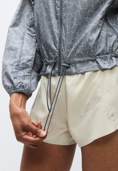 Γκρι ελαφρύ μπουφάν με σχέδιο και ελαστική ζώνη στη μέση, σε συνδυασμό με εκρού σορτς που διαθέτουν δίχτυ και ρυθμιζόμενα κορδόνια.