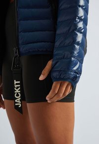 Gros plan sur une personne portant une veste matelassée bleue et un short noir, avec une sangle noire visible portant l'inscription "JACKIT" suspendue à la fermeture éclair de la veste.