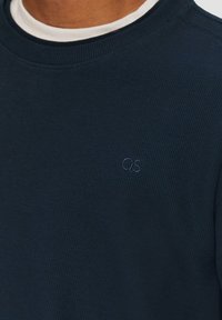 Marineblauwe gebreide sweater met een geribbelde textuur en subtiele QS-logo op de borst. Afgewerkt met een ronde hals en lange mouwen.