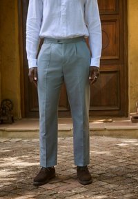 Hellblaue maßgeschneiderte Hose mit Falten, aus glattem Stoff, kombiniert mit braunen Wildlederschuhen. Trägt ein weißes Hemd mit Knöpfen.