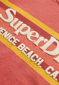 Nærbillede af tekstureret hvid skrift "SuperDry" og "VENICE BEACH. CA." på en pink, rød og gul stribet stofbaggrund.