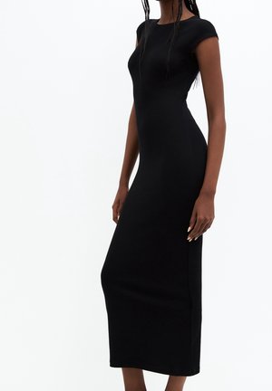 Femme portant une robe midi noire, ajustée, à manches courtes, debout devant un fond blanc uni.