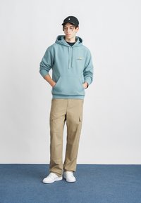 Felpa blu chiaro con tasca frontale e cordino, abbinata a pantaloni cargo beige con tasche laterali e vestibilità rilassata. Sneakers bianche.