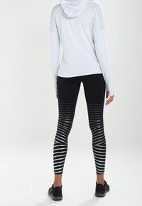 Maglietta bianca a maniche lunghe con cappuccio e leggings neri con strisce bianche orizzontali e verticali, abbinati a scarpe nere.