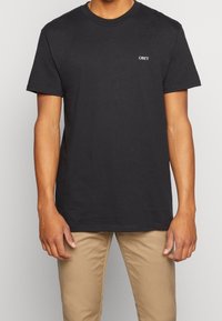 Svart bomulls-T-shirt med rund halsringning och korta ärmar, med en vit "OBEY"-logotyp på vänster bröst. Klassisk passform.