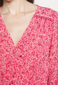 Blommig klänning i nyanser av rosa, med en djup V-halsringning, rynkade axlar och ett lätt, texturerat tyg med intrikata mönster.
