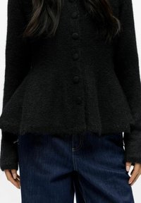 Musta neulottu neuletakki, jossa on peplum-muotoilu, teksturoitu kangas, napit edessä ja pitkät hihat, yhdistetty tummiin farkkuihin.