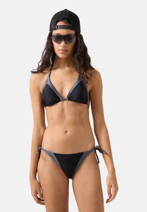 Kobieta ubrana w czarno-szary bikini z topem typu halter, czarną czapkę założoną tył na przód oraz ciemne okulary przeciwsłoneczne na jednolitym jasnym tle.