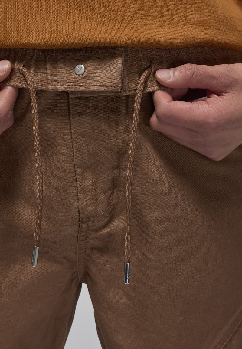 Pantaloncini marroni con vita regolabile tramite cordino, dotati di dettagli in metallo argentato e una superficie liscia, mostrati mentre vengono tenuti in mano per una regolazione.