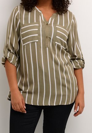 Olivgrüne Bluse mit langen Ärmeln, weißen vertikalen Streifen und zwei Brusttaschen, getragen zu schwarzer Hose. Ärmel bis zu den Ellbogen hochgekrempelt.