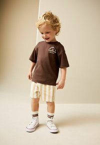 Braunes T-Shirt mit kurzen Ärmeln und weißem Logo, gelb-weiß gestreifte Shorts, weiße Sneaker mit roten Akzenten und weiß-braun gestreifte Socken.