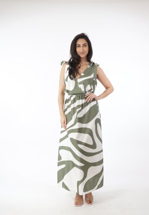 Mujer con cabello largo y oscuro que lleva un vestido maxi sin mangas con un patrón abstracto verde y blanco, de pie con una mano en la cadera.