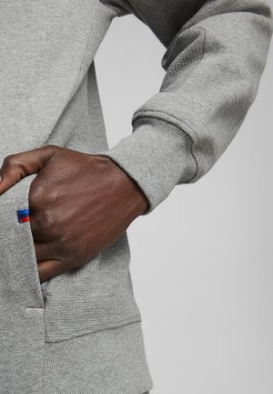 Szary sweatshirt z ściągaczami przy rękawach, z ręką w kieszeni. Zauważalnym detalem jest mała czerwona i niebieska metka przy krawędzi kieszeni.