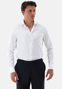 Camicia bianca a maniche lunghe con bottoni e colletto classico, abbinata a pantaloni scuri. La camicia ha una texture liscia e una vestibilità regolare.