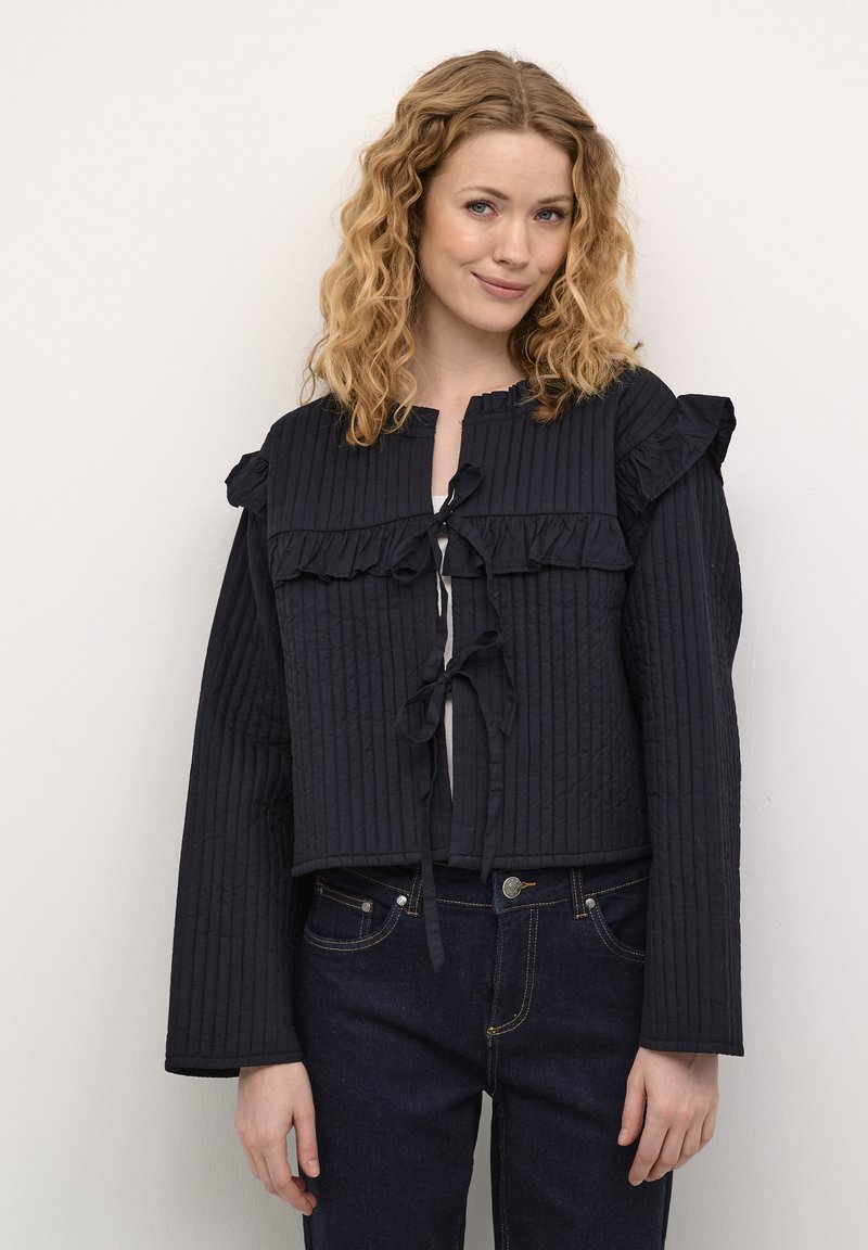 Veste noire matelassée avec des accents volants, des manches longues et un système de fermeture à nouer à l'avant. Portée au-dessus d'un haut clair et associée à un jean foncé.