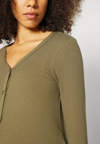 Top de manga longa em verde oliva com texturização canelada, apresentando um decote em V e detalhes em botão, com uma textura suave e design ajustado.