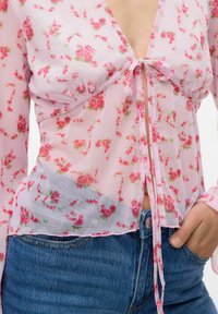 Blouse en voile floral rose avec un décolleté plongeant, un nœud devant et des manches longues. Présente de petits motifs de roses et une texture légèrement froncée.
