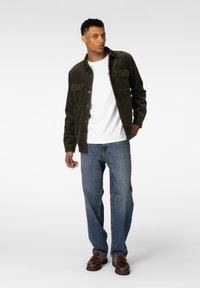 Mørkegrønn kordfløyelsjakke med knappelukking, to brystlommer, hvit t-skjorte under, blå jeans og brune snøresko.