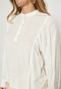 Balta, tekstūruota bliuskutė su stačiu apykakle, mygtukais priekyje ir raukiančiomis rankovėmis. Yra subtilus languotas raštas ir šoninės plyšiai.