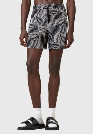 Sorte og hvide shorts med palmebladmønster, udstyret med elastisk talje, afslappet pasform og længde til midten af låret. Stylede med sorte sandaler og hvide sokker.