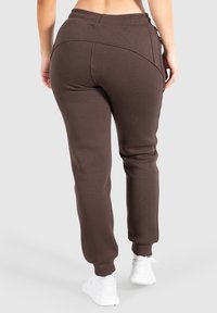 Rückansicht einer Person, die braune Jogginghosen und weiße Sneakers vor einem schlichten Hintergrund trägt.