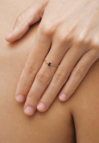 Anillo de oro con una banda delgada, que presenta una pequeña piedra negra rectangular y una diminuta piedra clara de acento. La mano descansa sobre una superficie de piel bronceada.