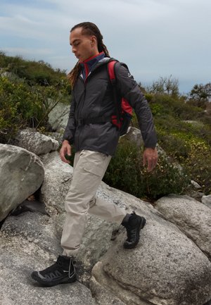 Veste de plein air gris foncé avec des accents rouges, pantalon beige et bottes de randonnée noires. Terrain accidenté avec des rochers et de la végétation visibles.