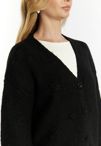 Zwarte gebreide cardigan met een V-hals, voorzien van gestructureerde bloempatronen, lange mouwen en een dubbele knoopsluiting aan de voorkant.