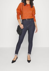 Pomarańczowy wełniany sweter, ciemnoniebieskie obcisłe dżinsy, czarny pasek i czarna pleciona torebka; modelka nosi czarne spiczaste baleriny na niskim obcasie.